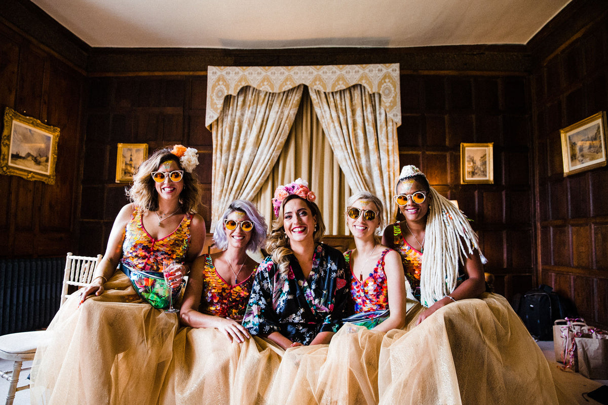 A bride wearing a festival style headdress sits with her bridesmaids on the morning of her wedding and they are all wearing sun glasses and pink sequin playsuits. 