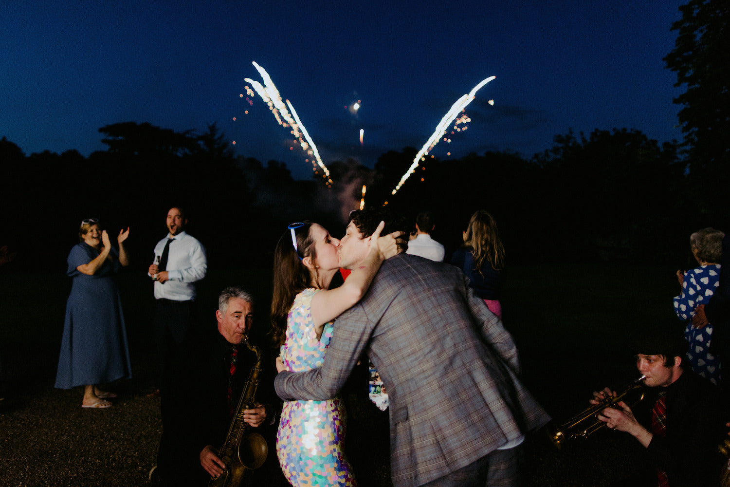 A bride wearing a white sequin jumpsuit kisses the groom while fireworks go off in the background. 