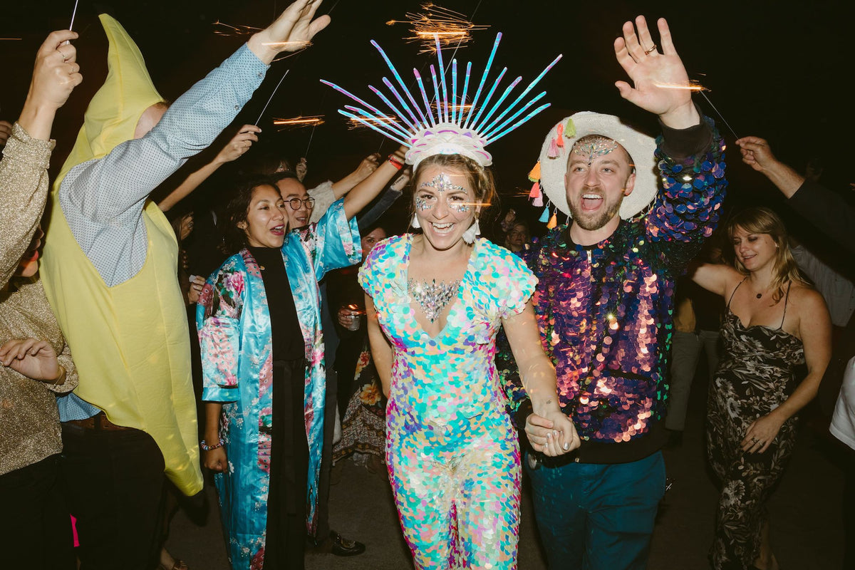 A festival style bride wearing an ornate headdress and white sequin jumpsuit holds hands with her partner who is also wearing a sequin bomber jacket with a cowboy hat. 