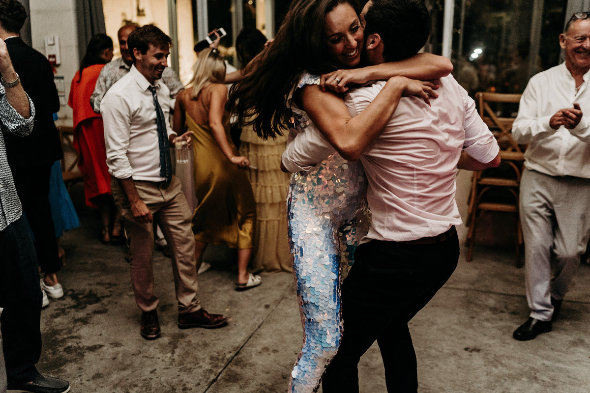 A bride wearing a white sequin jumpsuit dances with her husband