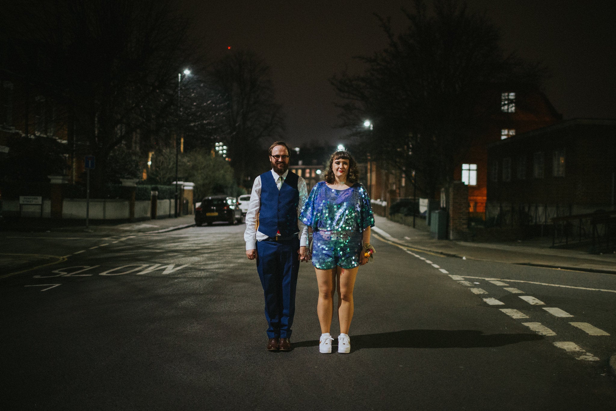 A bride wearing a blue sequin romper stands with her husband at night in a city street. 
