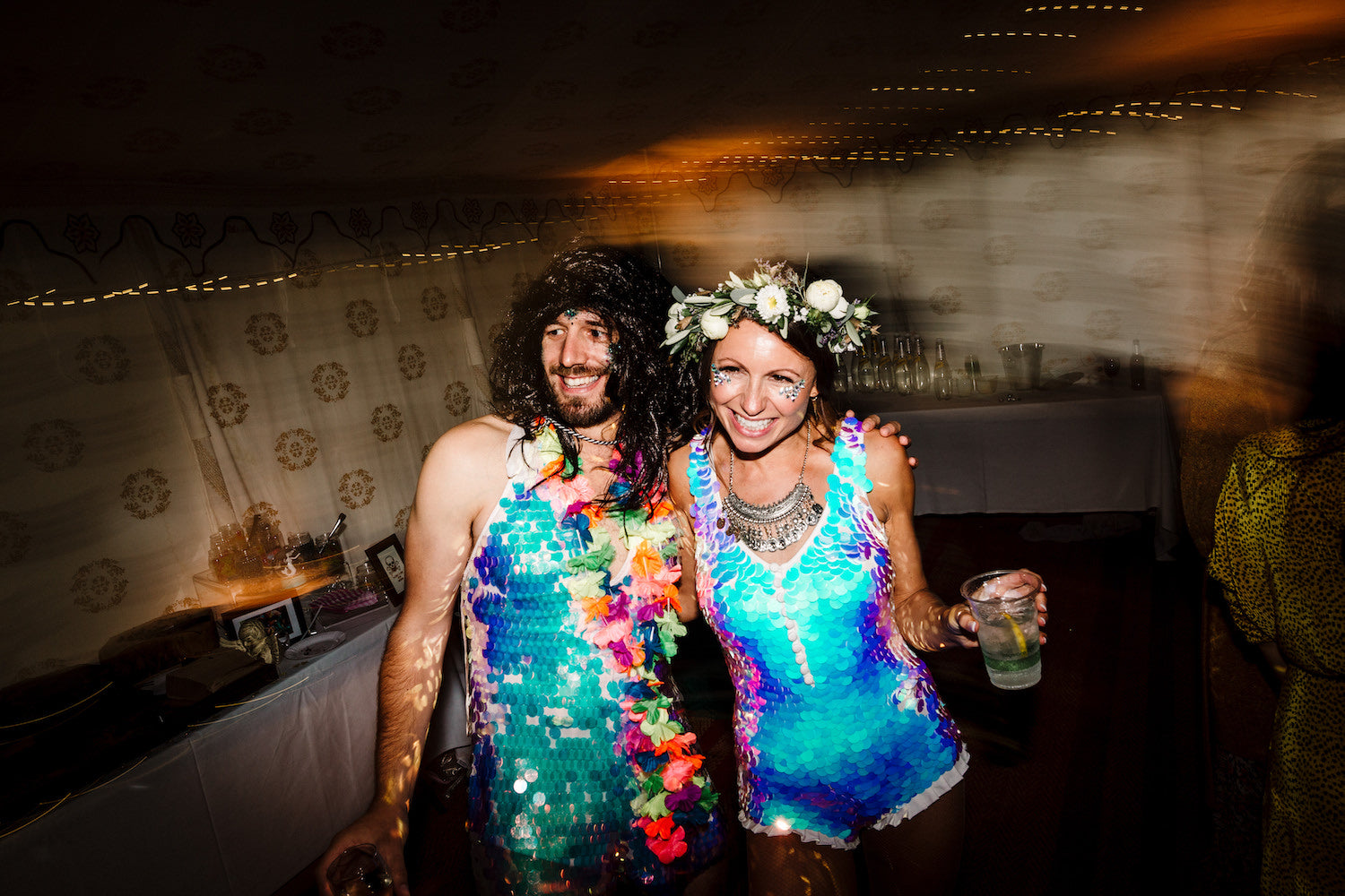 A bride wearing a white sequin playsuit poses with a male wedding guest wearing a sequin dress and a long black wig.