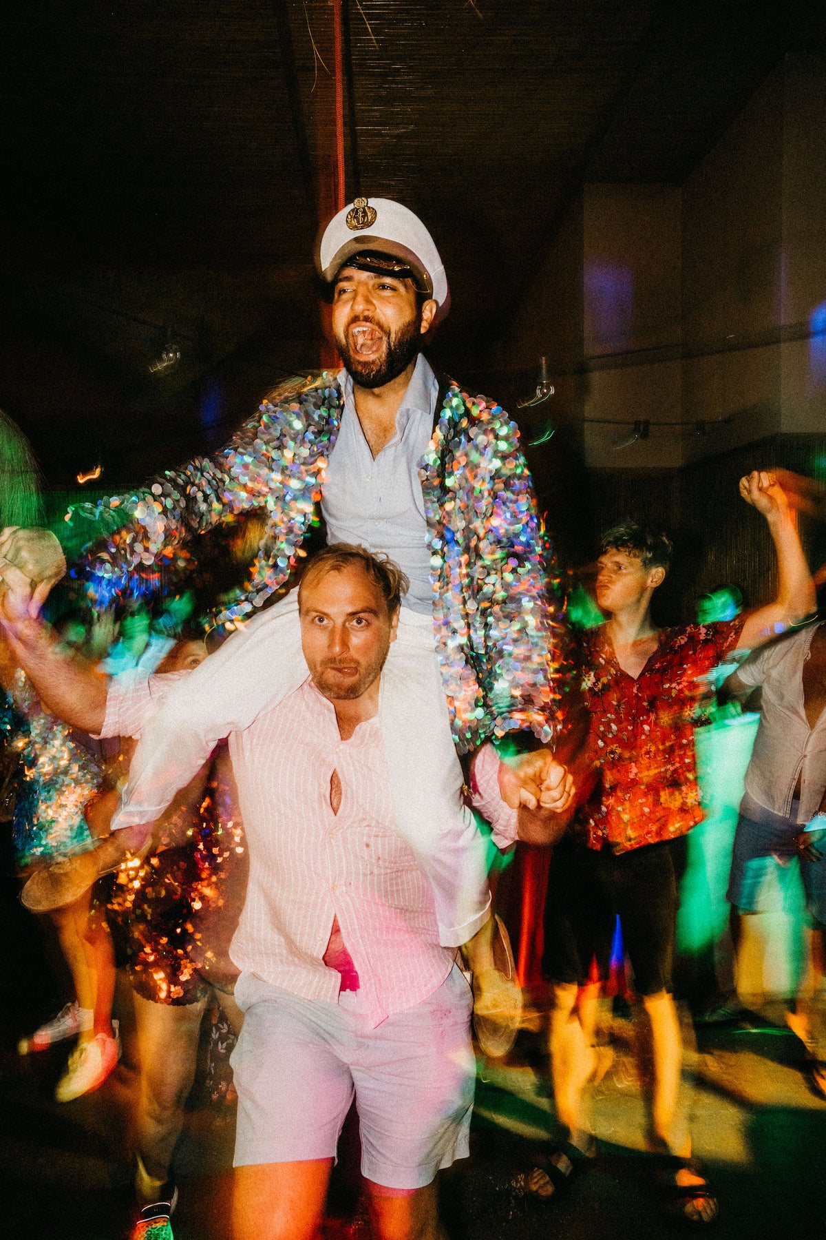 A groom wearing a festival style sequin bomber jacket  and sailor hat sits on the shoulders of his friend at his wedding. 