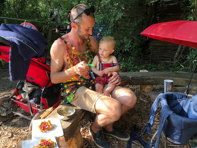 Sam Bloom wearing a coral and aqua sequin vest sitting on a bench at Glastonbury Festival feeding his baby son