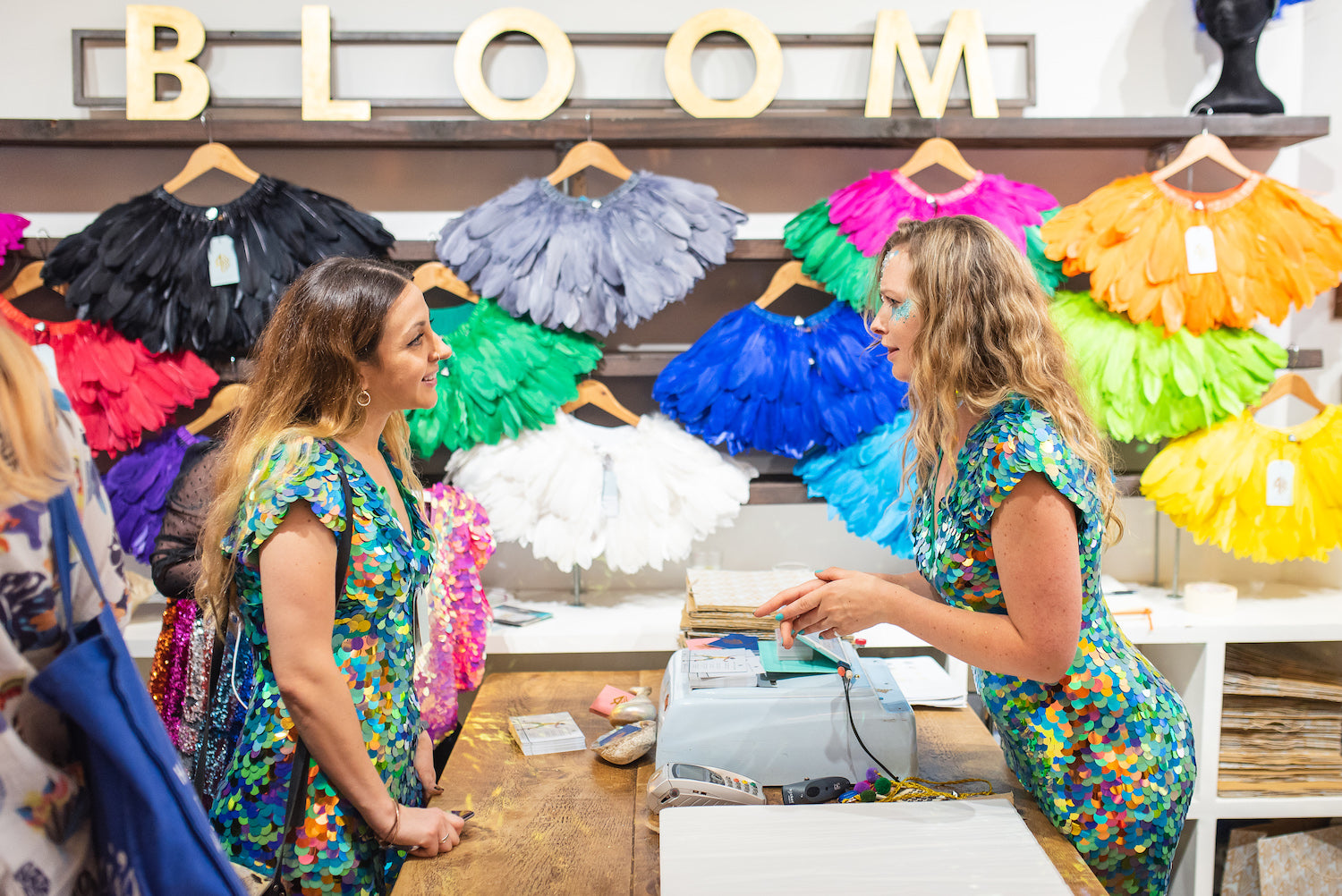 Two women wearing green sequin jumpsuits by slow fashion brand Rosa Bloom talk over the counter at the Rosa Bloom Pop Up Shop. 