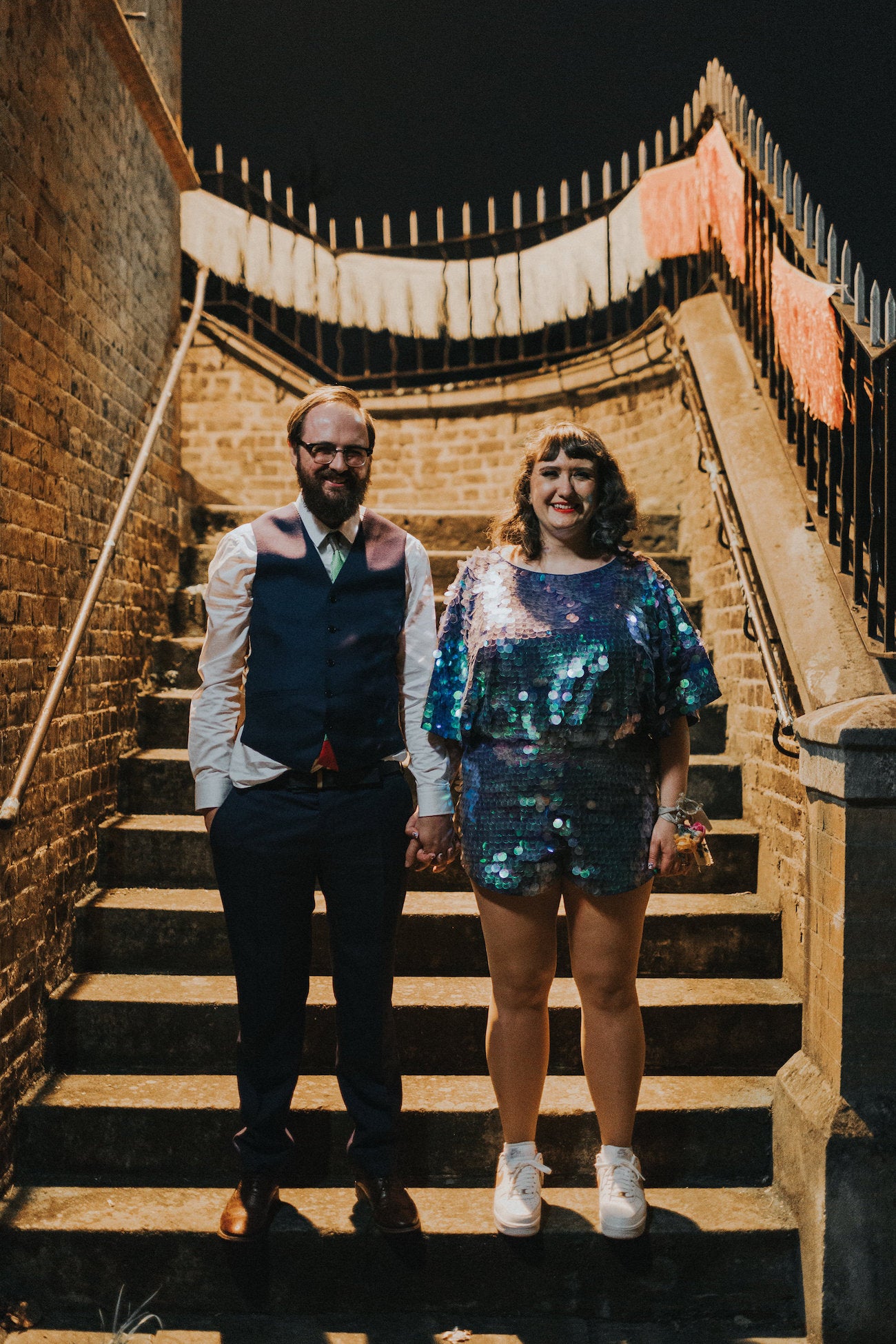 A newly wed couple pose for a photograph at night by some stone stairs and the woman wears a blue sequin romper. 