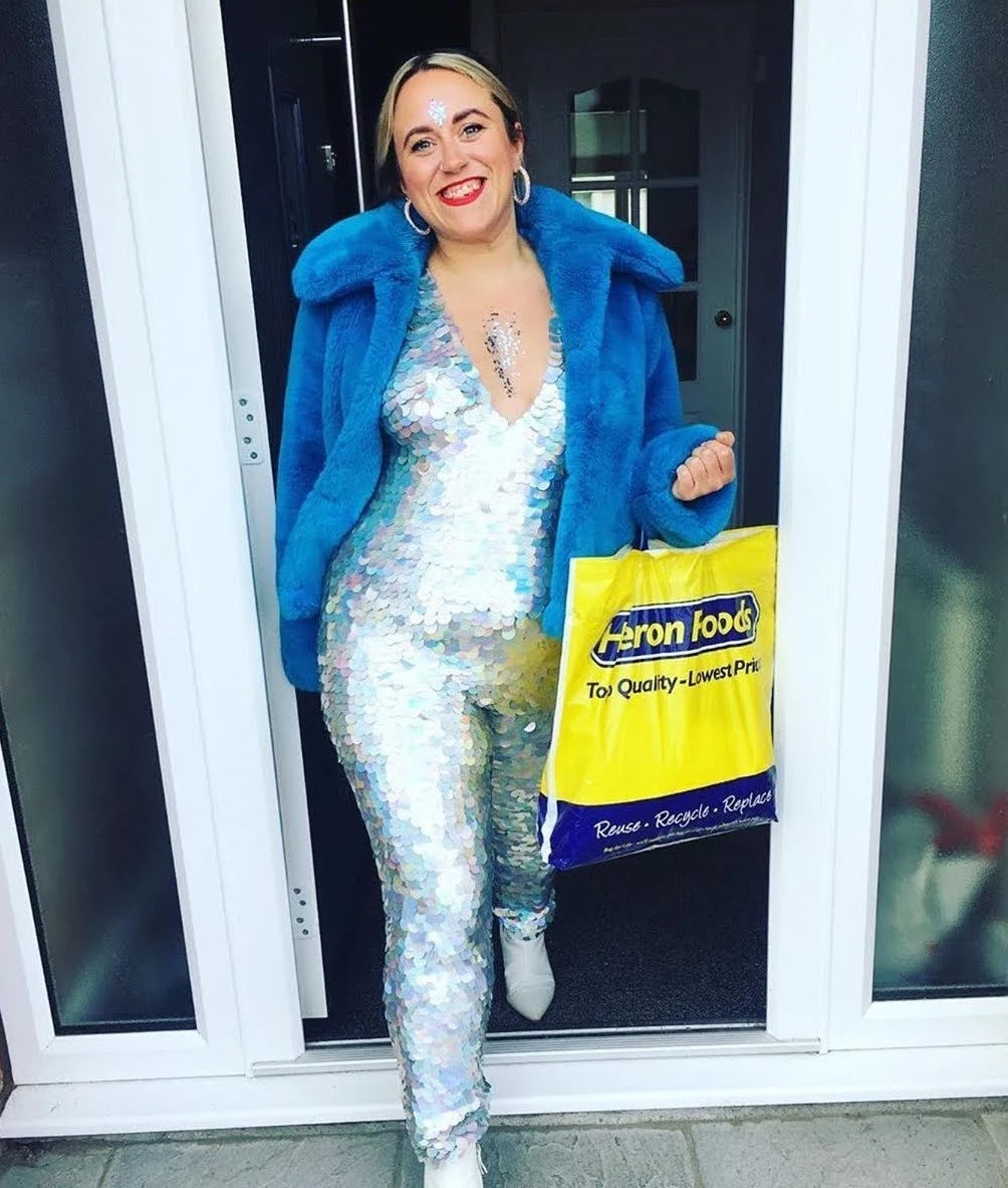 woman stepping out of a supermarket wearing a silver sequin jumpsuit and blue jacket