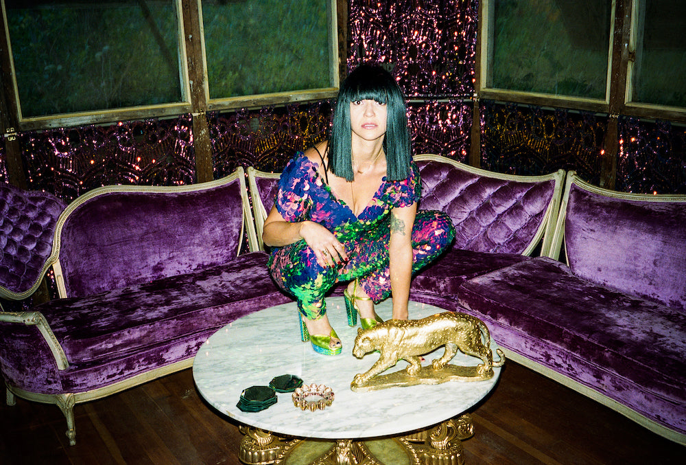 Laura Leezy crouching on a marble coffee table wearing a tight Rosa Bloom jumpsuit covered in large round iridescent purple sequins and metallic heels in front of lavish purple velvet couches and a gold tiger ornament on the table