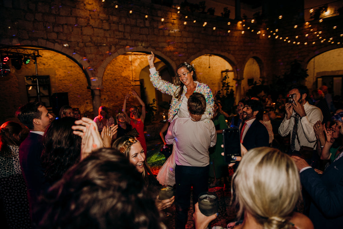 A bride wearing a festival style sequin bomber jacket and glitter makeup is held up by her husband on the dancefloor 