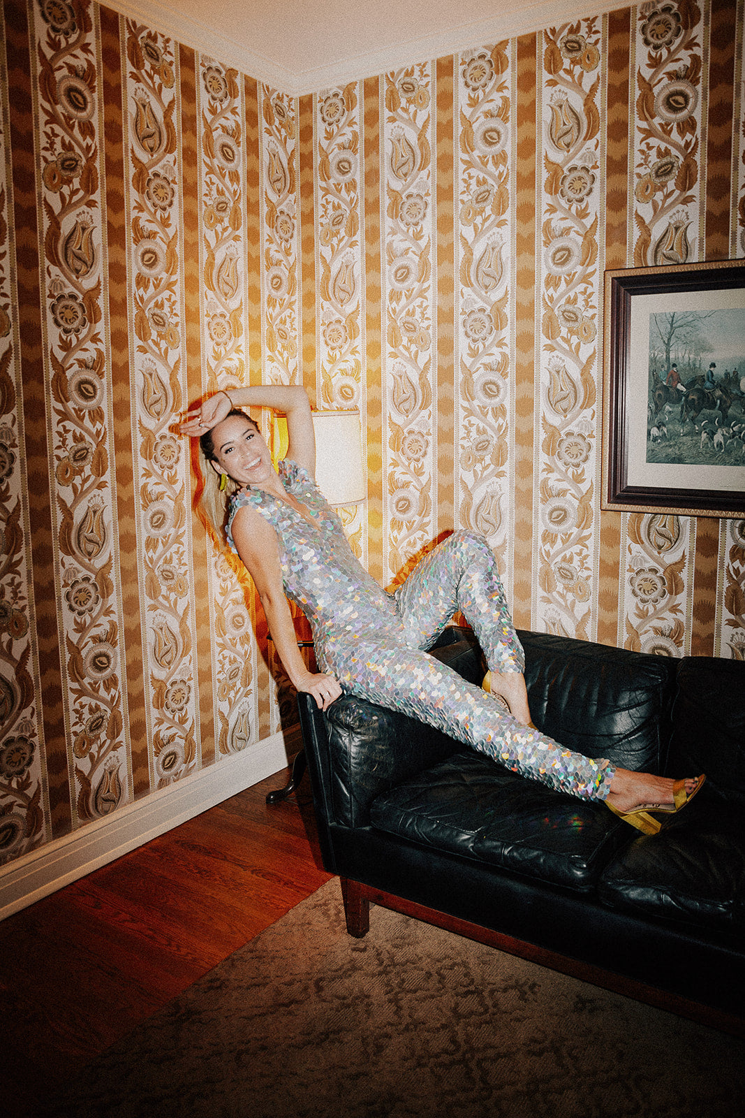 A bride wearing a festival style silver sequin jumpsuit strikes a pose across a sofa at her wedding. 