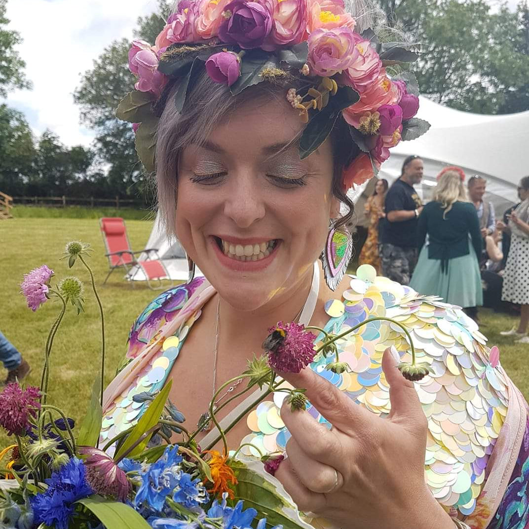 A bride wearing a festival sequin jumpsuit and headdress admires a bee in her bouquet.