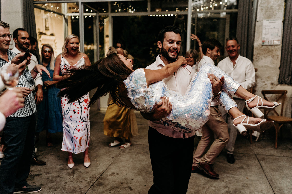 A groom carries his bride who is wearing a white sequin jumpsuit