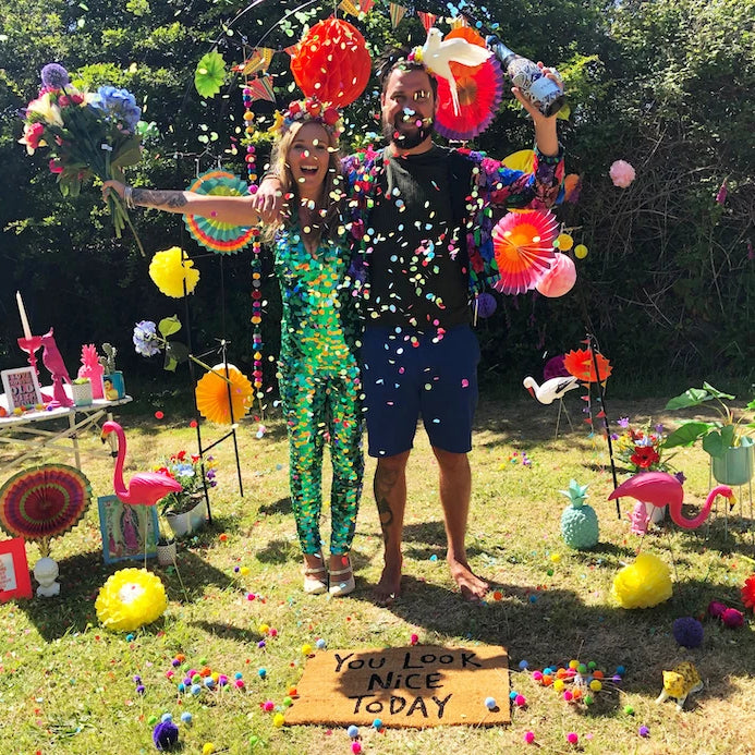 Man and woman stood in a garden filled with colourful props throw confetti