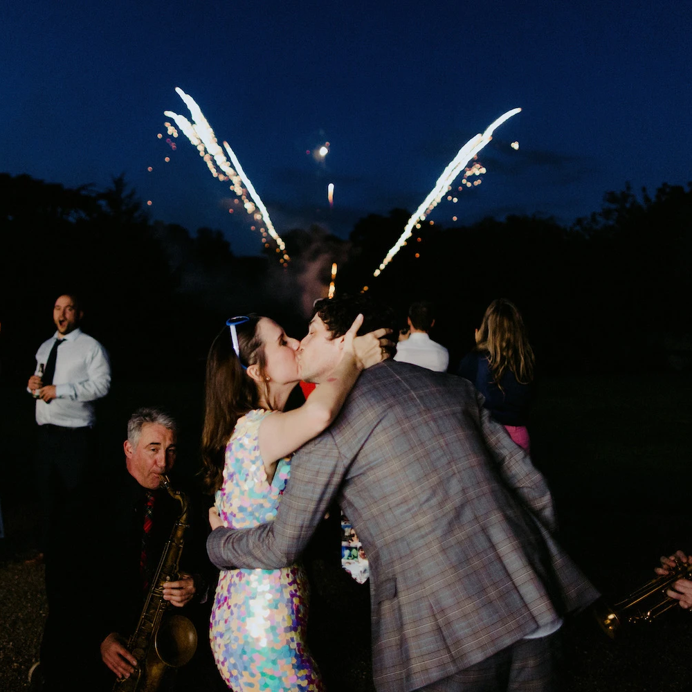 A bride wearing a festival style sequin jumpsuit kises her husband on their wedding day while fireworks go off in the background