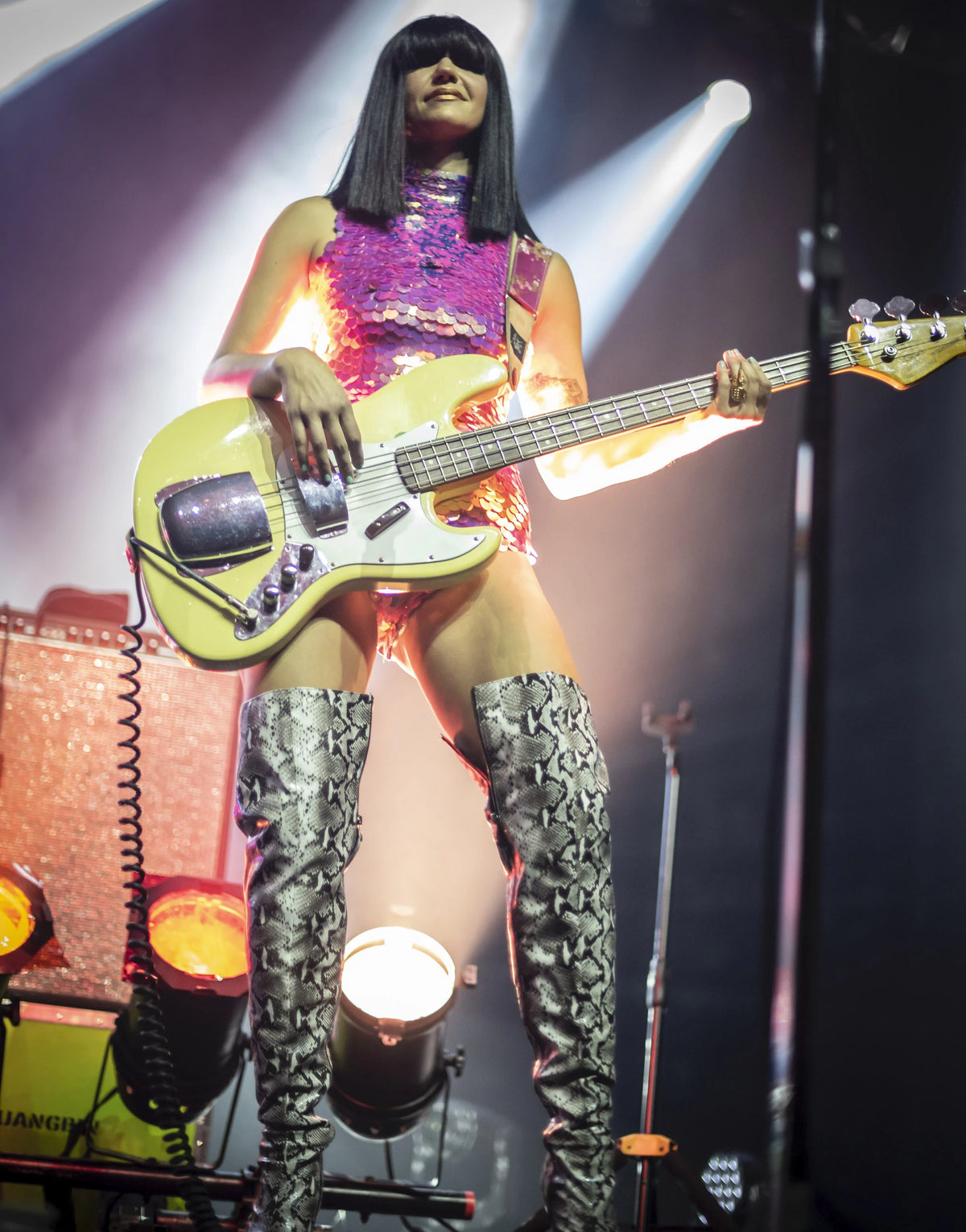 Laura Leezy on stage playing the bass guitar wearing thigh high snake skin boots and high neck leotard covered in large round iridescent hot pink sequins