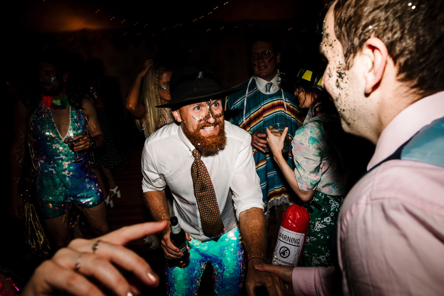 A groom wearing white sequin leggings a white shirt and a black hat is dancing with friends at his wedding reception 