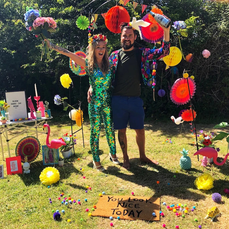 Man and woman stand in a garden surrounded by colourful props and the woman is wearung a green sequin jumpsuit