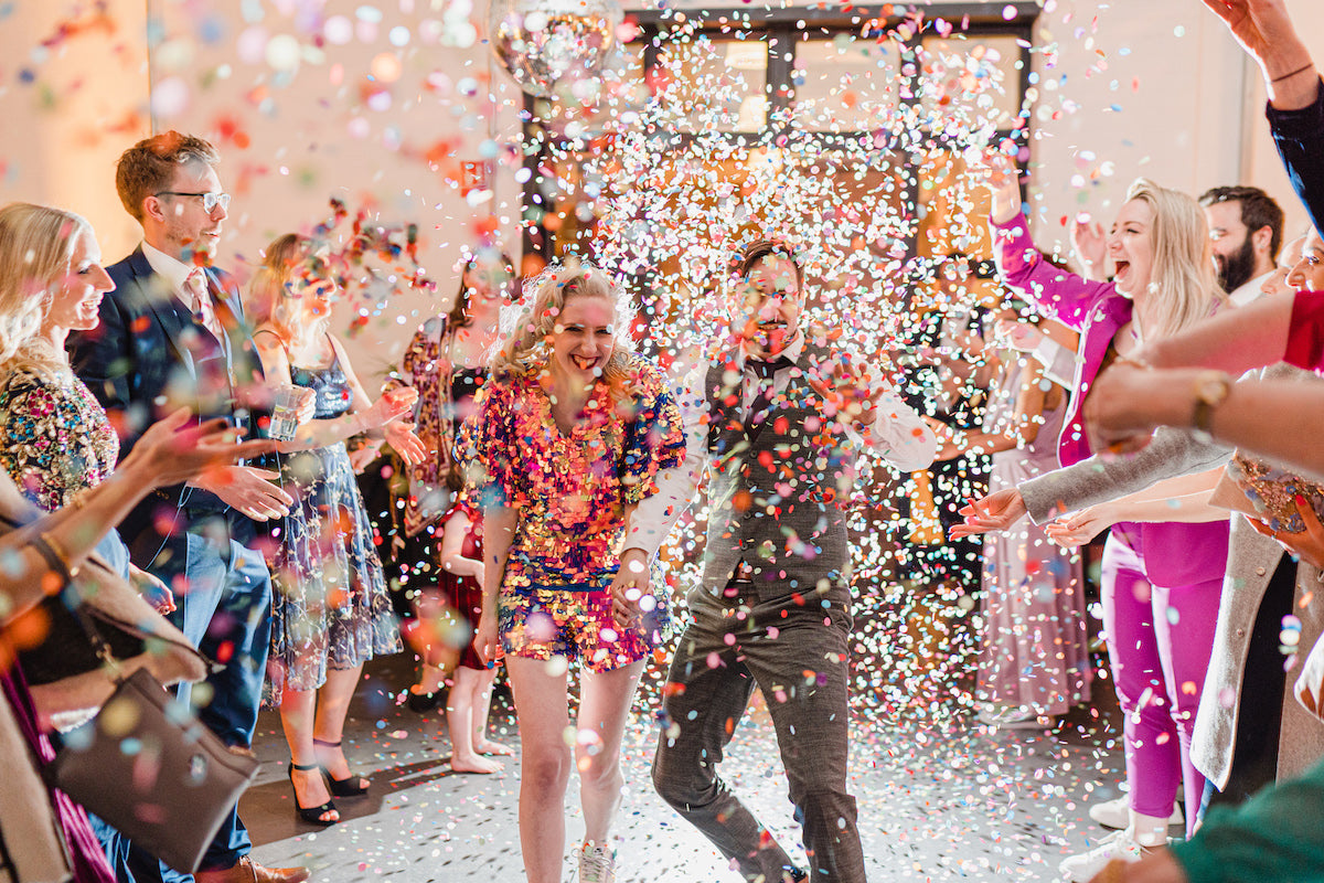 A bride wearing a purple sequin playsuit and a groom are showered with colourful confetti