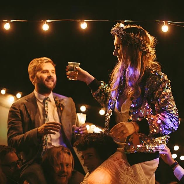 A bride and groom sit on the shoulders of their guests, the bride is wearing a festival style floral crown and sequin bomber jacket.