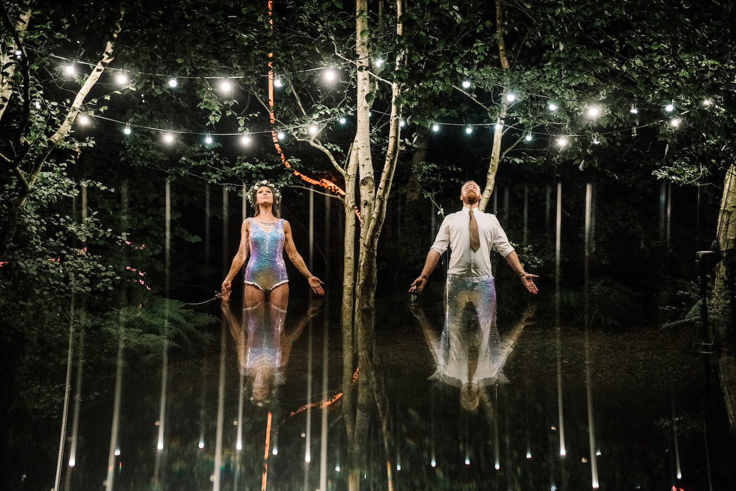 Bride and Groom wearing matching white sequin outfits stand with their arms open under fairy lights.