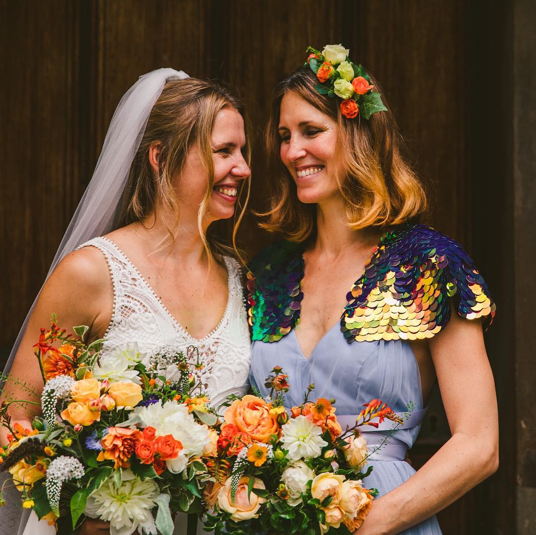 A bride stands with her bridesmaid who is wearing a purple sequin cape and a festival style floral headdress.
