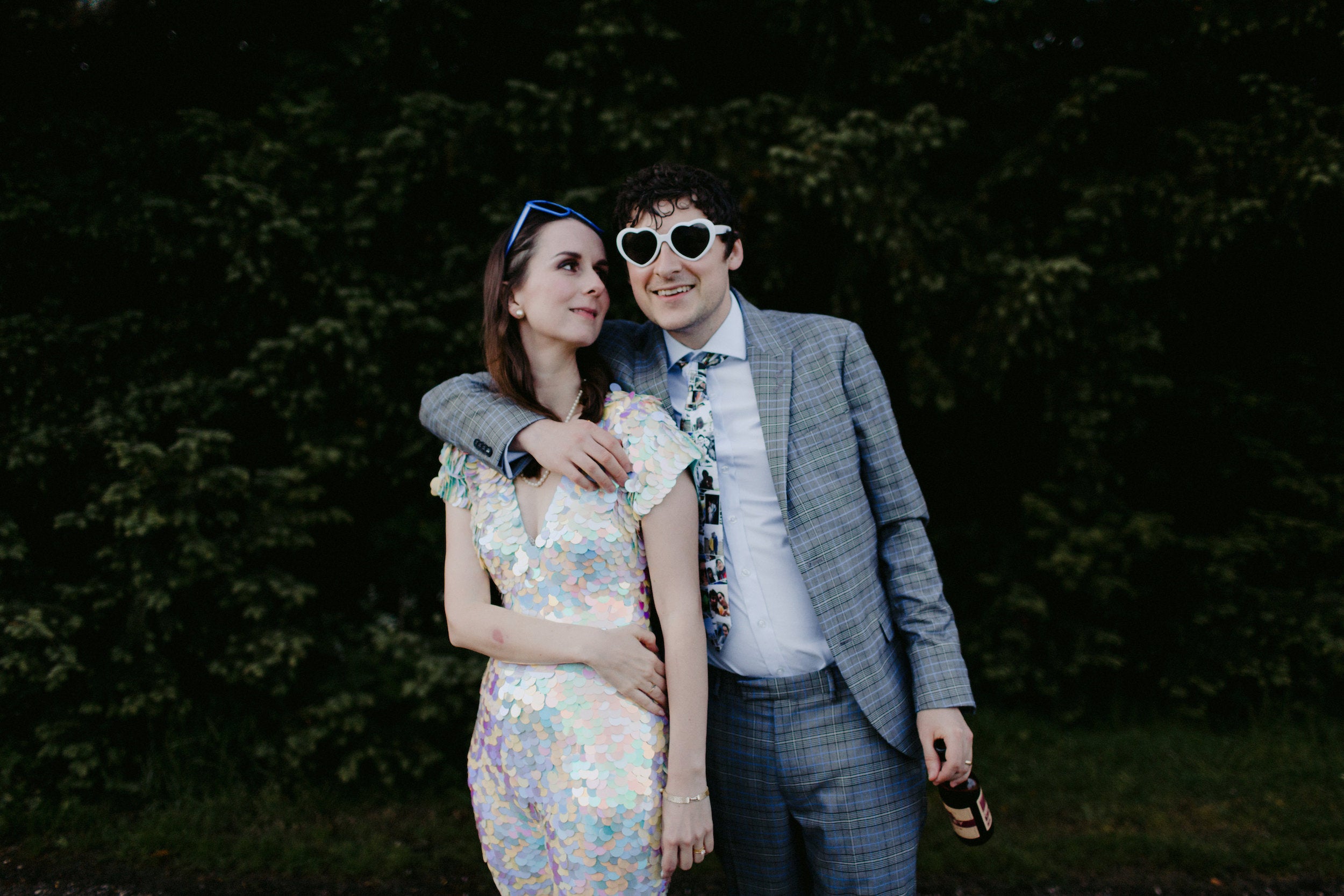 Bride wearing a white sequin outfit stands with the groom who wear a suit and heart shaped sunglasses.