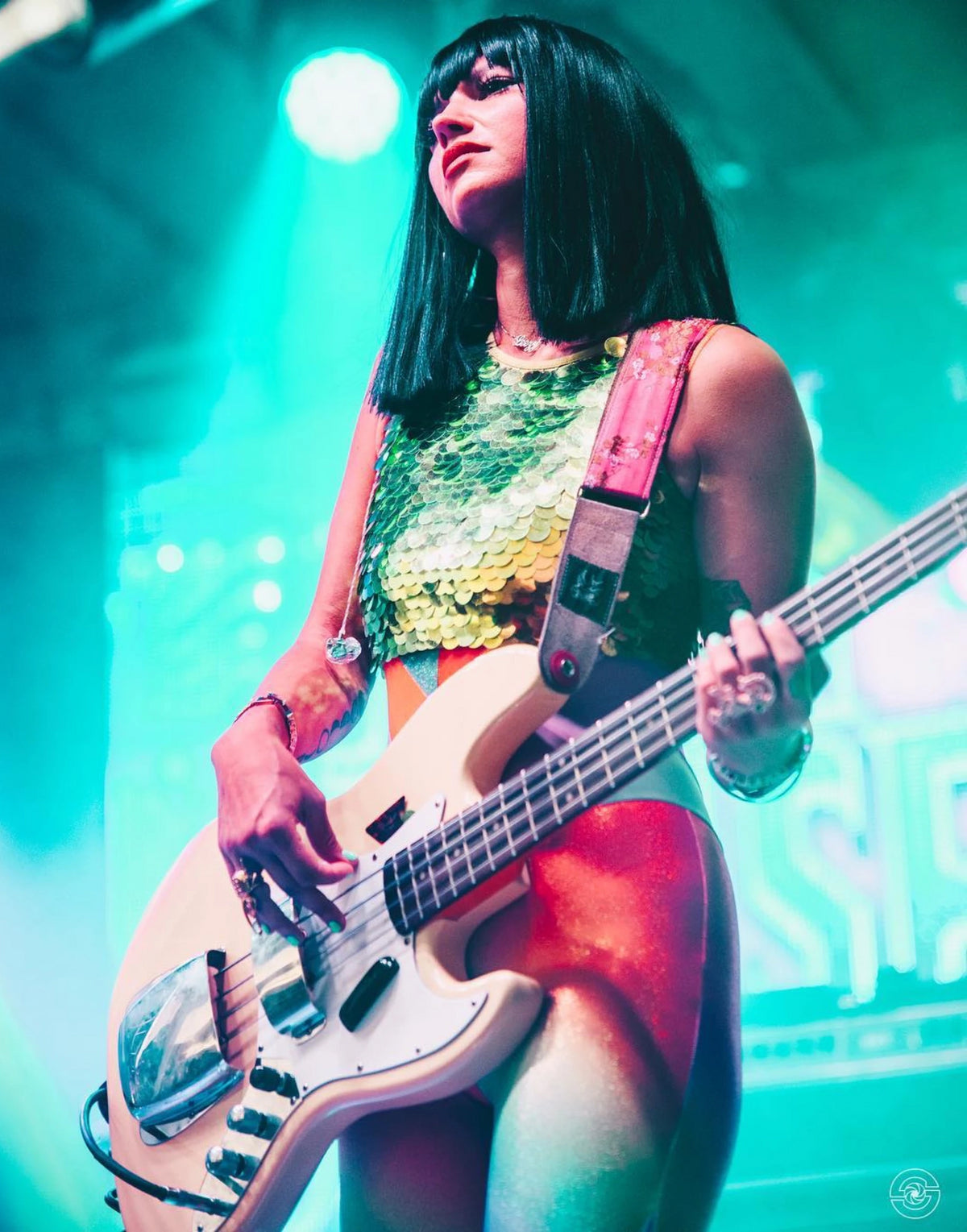 Laura Leezy on stage playing the bass guitar wearing a black Cleopatra style wig and a green sequin Rosa Bloom Twinks crop top over a lycra catsuit with aqua lighting behind