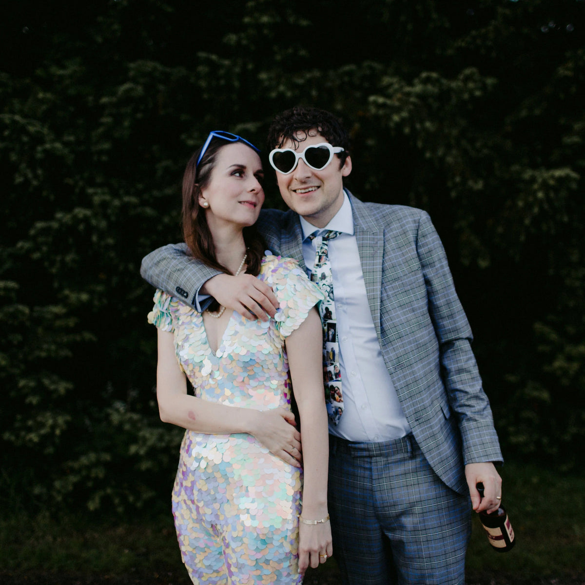 A bride wearing a festival style white sequin jumpsuit poses with her husband who is wearing a suit and sunglasses