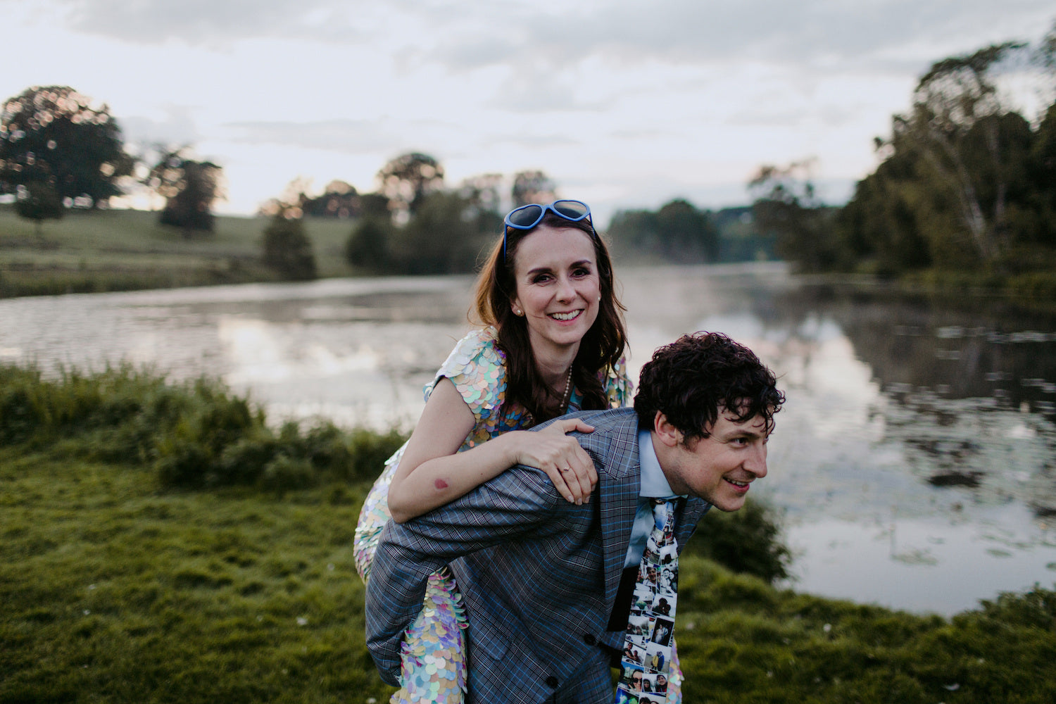 A bride wearing a white sequin jumpsuit and heart shaped sun-glasses gets a piggy-back form her groom. 