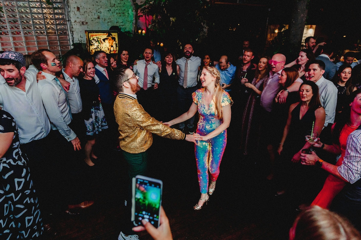A bride and groom dance together at their wedding reception and both are wearing bright sequin outfits.