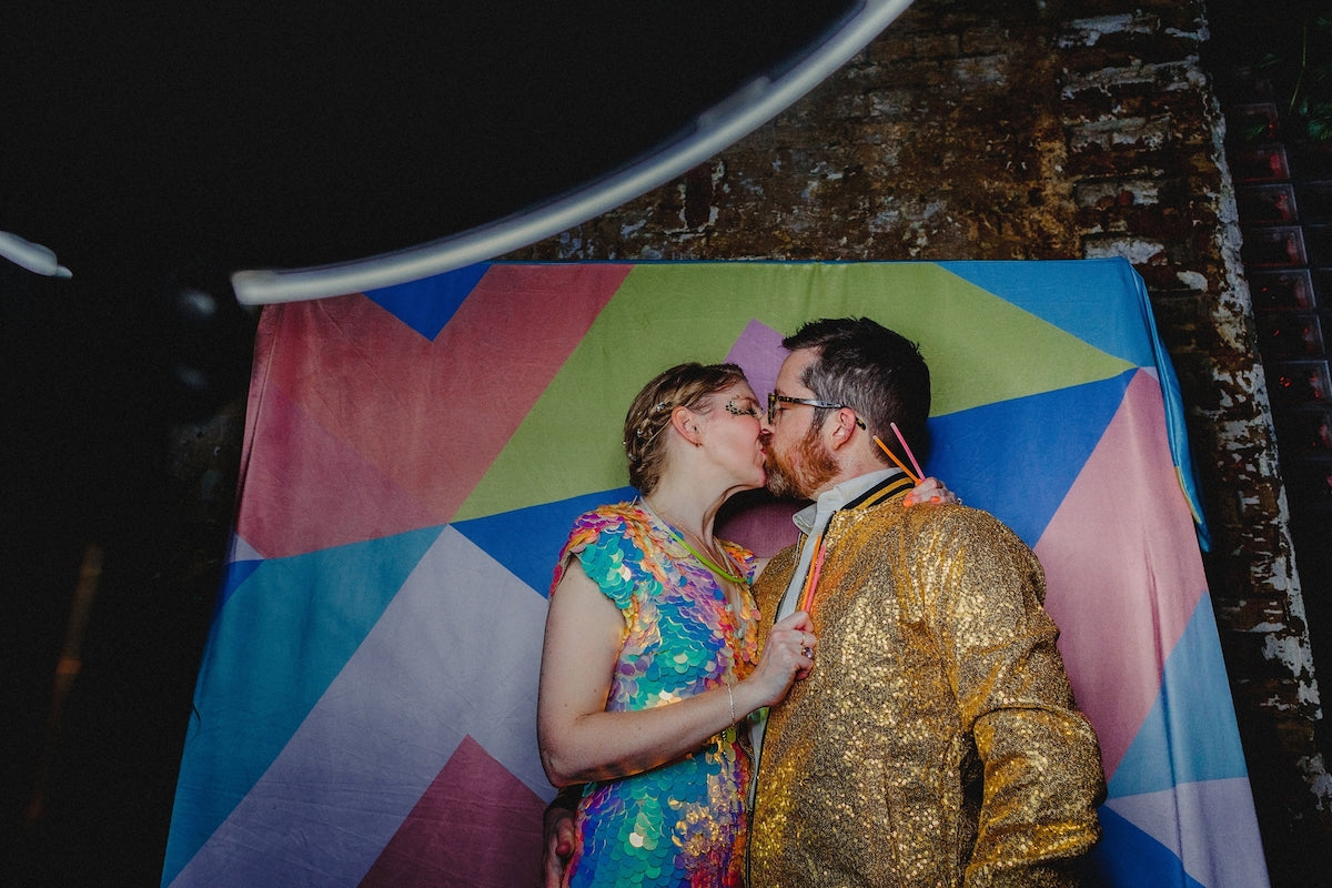 A bride wearing a rose pink jumpsuit kisses her husband in front of a bright coloured wall-hanging. 