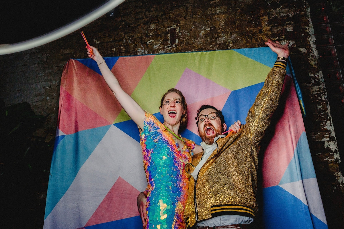 A bride and groom wearing bright sequin outfits pose together with their arms up in front of a bright coloured flag.