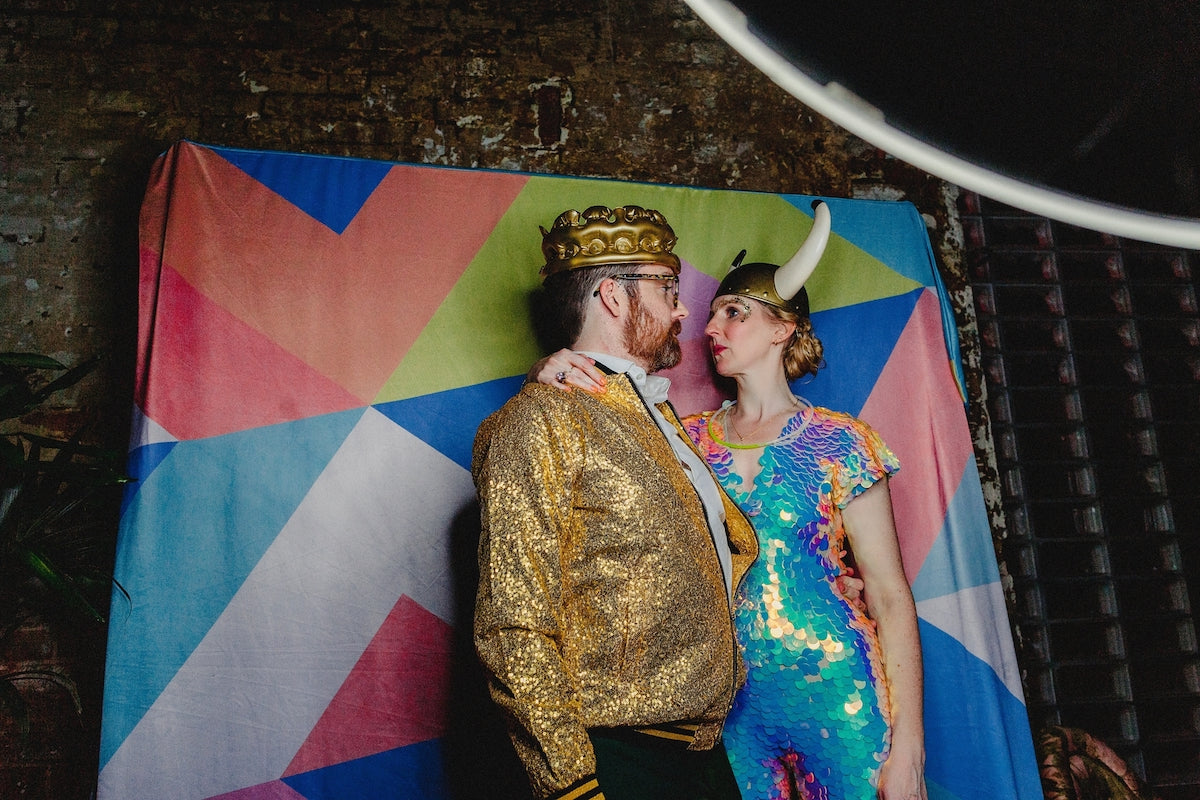 A bride wearing a rose sequin jumpsuit and a plastic viking party hat poses with her husband who also wears a gold sequin jacket and a plastic crown.