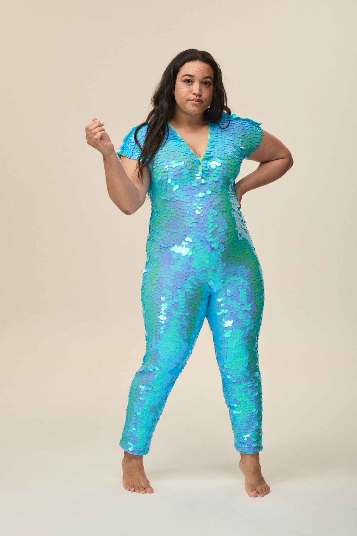 A plus sized woman wearing a green and blue sequin catsuit poses for the camera.