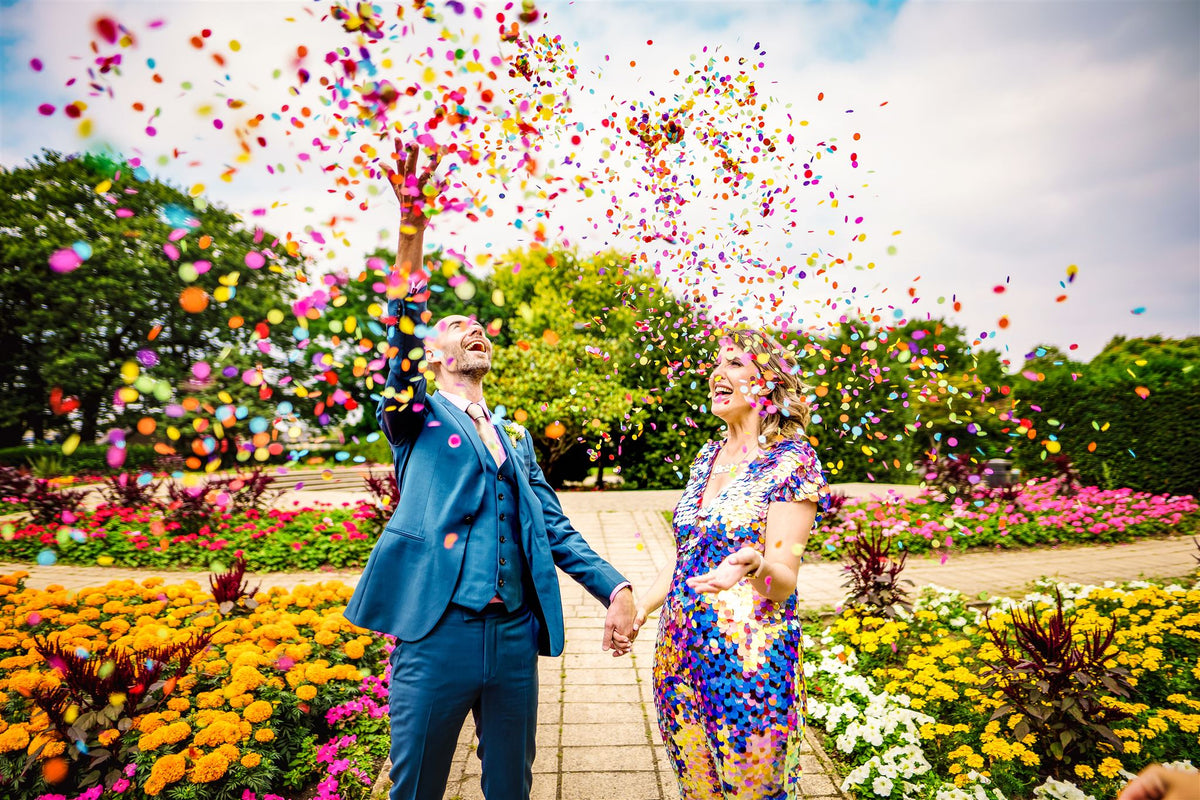 A bride wearing a purple sequin jumpsuit and  her groom are captured under a colourful cloud of confetti.