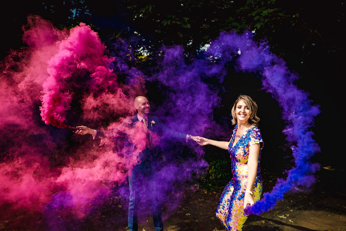 A bride wearing a purple sequin jumpsuit and her husband hold pink and purple smoke cannons.