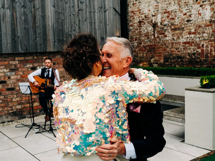 A bride wearing an off-white sequin bomber jacket hungs her father on her wedding day