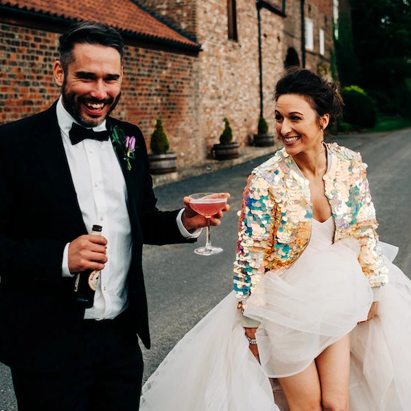 A bride wearing a cream sequin bomber jacket walks alongside her husband who is holding a glass of pink champagne.