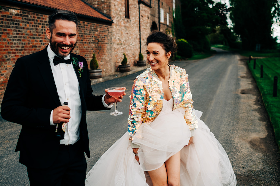 A bride wearing a cream sequin bomber jacket over her princess wedding dress walks next to her groom who is holding a glass of pink champagne.