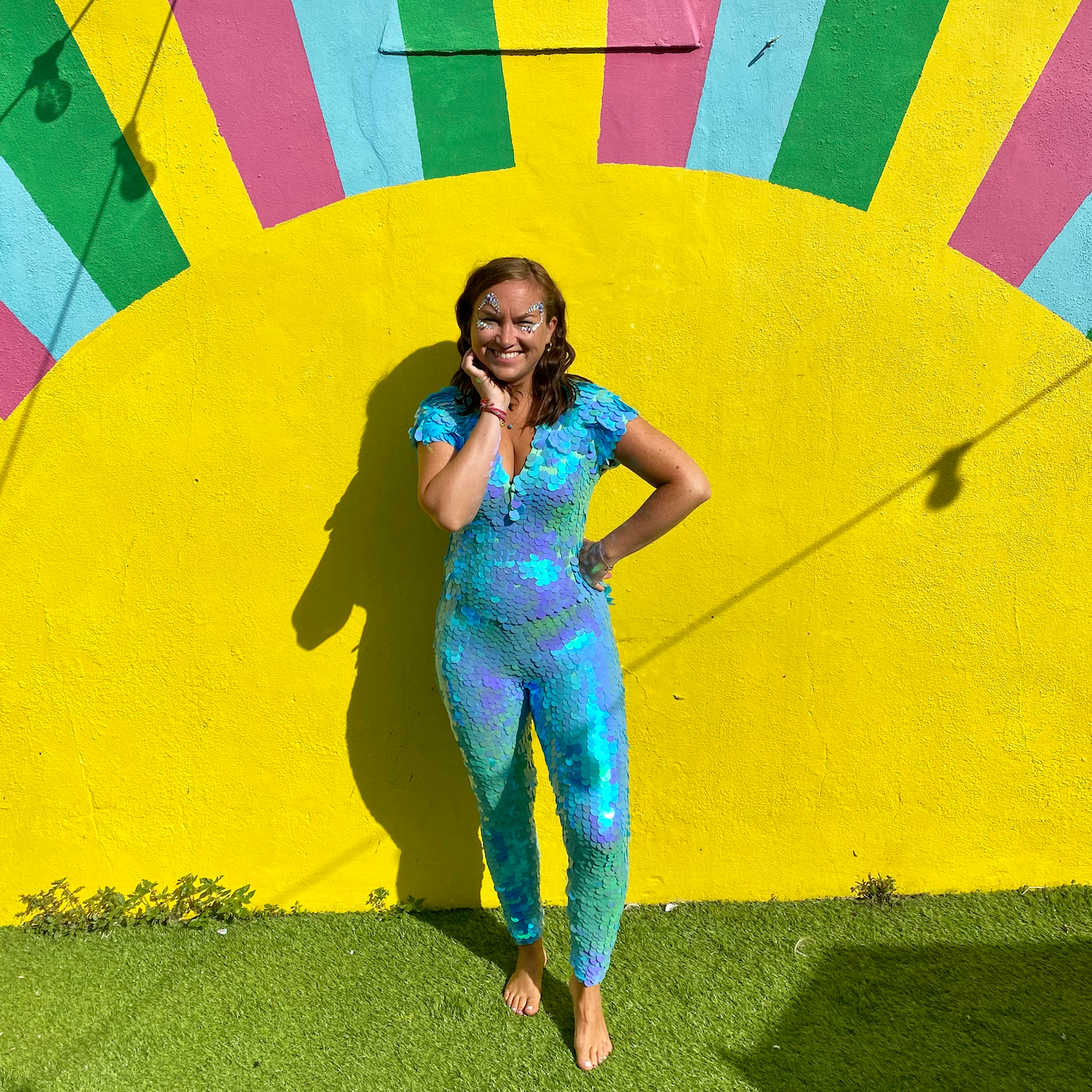 Smiling, petite woman stood in front of bright coloured sunshine wall wearing a Rosa Bloom Mint Aphrodite Sequin Jumpsuit