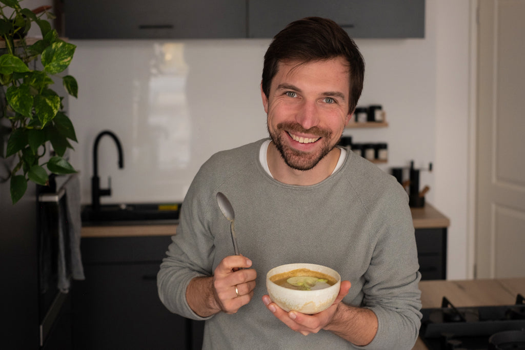Gründer Richard lächelnd mit einer Schüssel cremiger Maronen-Lauch-Suppe mit Miriquidi und Muskatnuss in der Hand und einem Löffel in der anderen 
