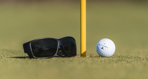 A pair of black Fuse Egmont sunglasses with Carbon AMP lenses placed next to a golf ball