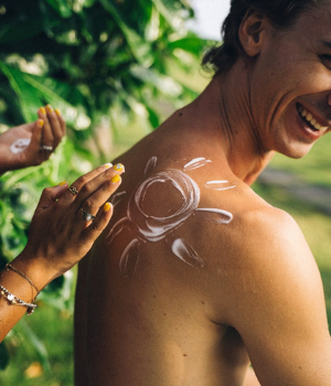 Man smiles as hands apply sunscreen to his back in the shape of a smiley face