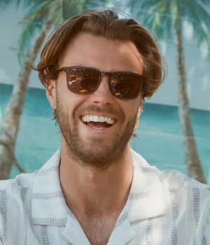 Man smiles with brown sumglasses on. Two palm trees in background