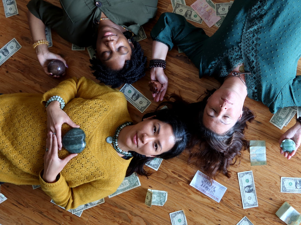 three women lying on the floor surrounded by  money holding abundance crystals