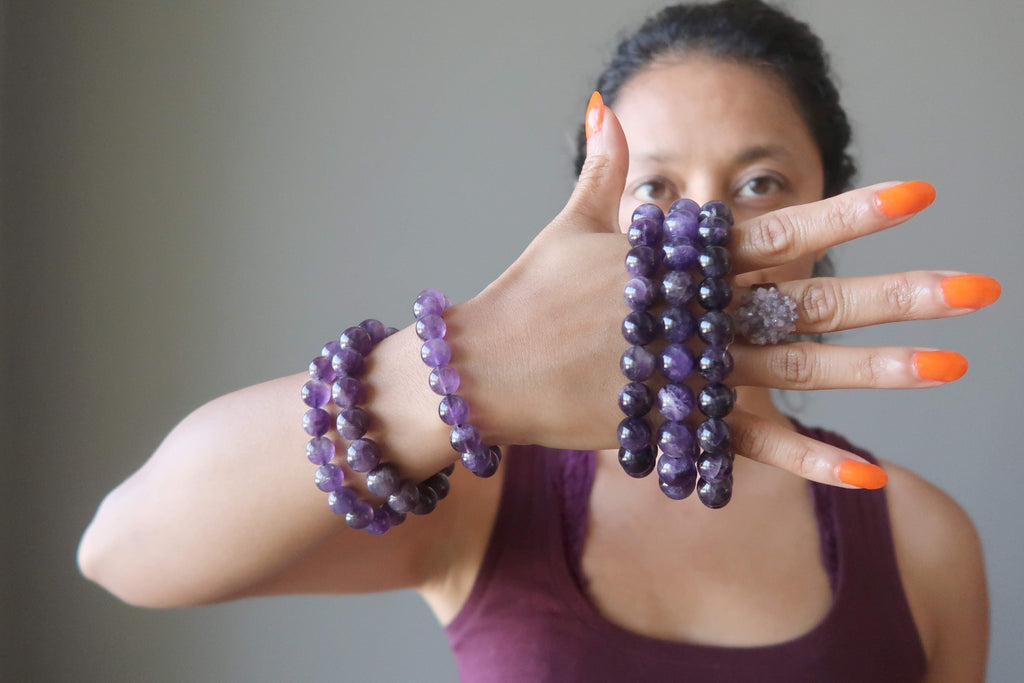 Model with hand full of Amethyst stretch bracelets  and Amethyst cluster ring standing against gray wall.