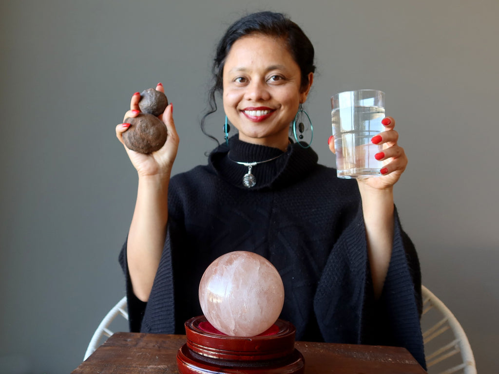 sheila of satin crystals sitting behind a red quartz sphere holding up moqui marbles and a glass of water for grounding