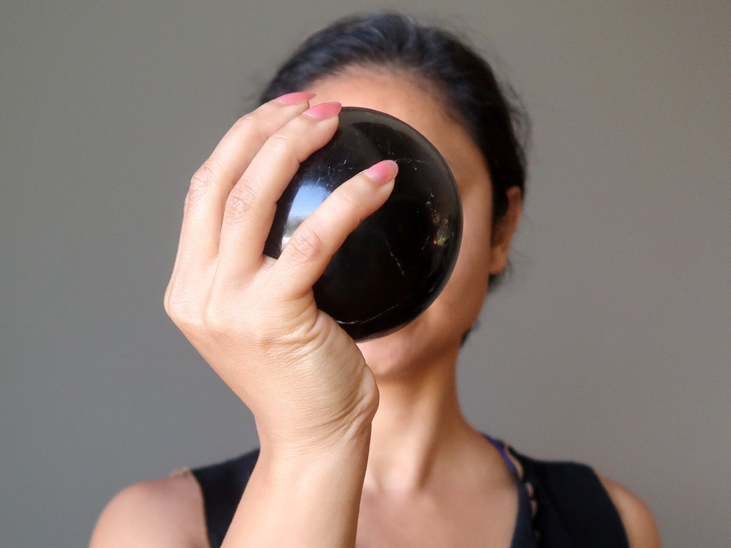 sheila from satin crystals holding up a black tourmaline stone sphere in front of her face