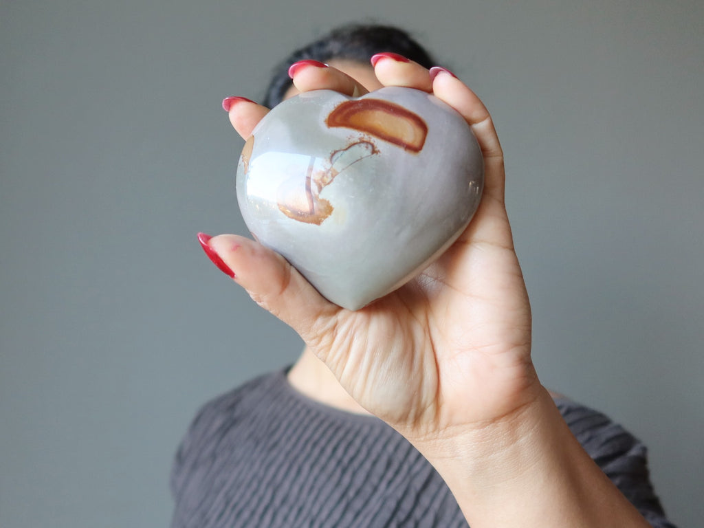 female holding a gray and orange polychrome jasper heart stone in front of her face