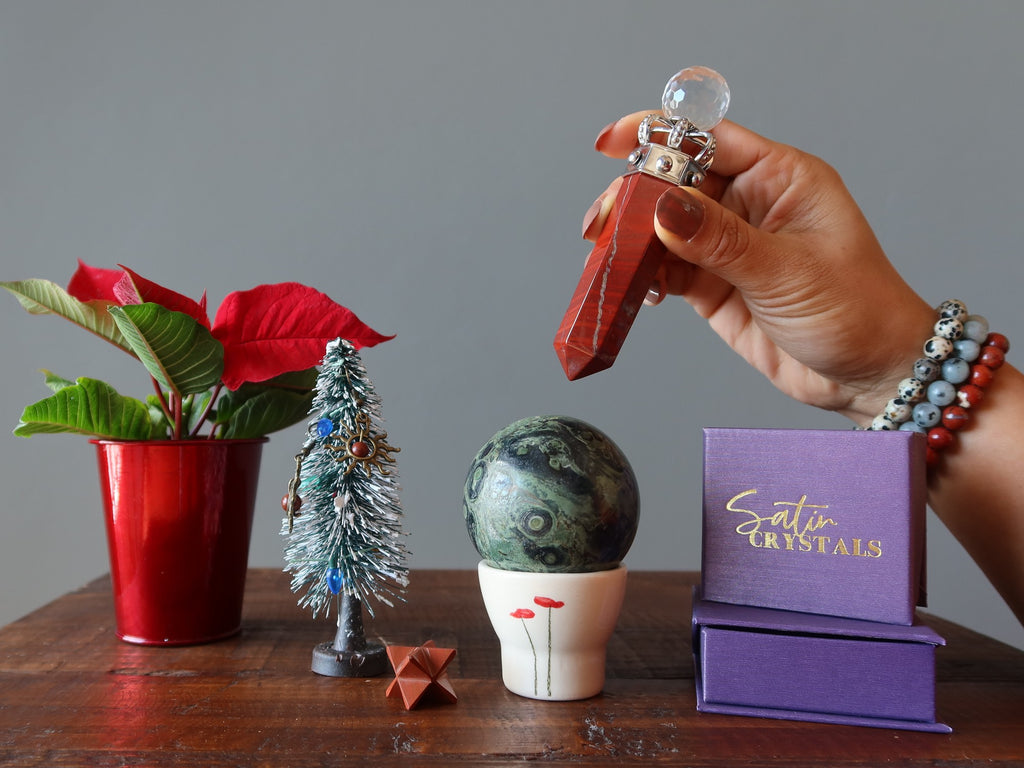 hand wearing jasper bracelets waving a red jasper wand over a kambaba jasper sphere on a table with red jasper merkaba, christmas tree, satin crystals gift boxes